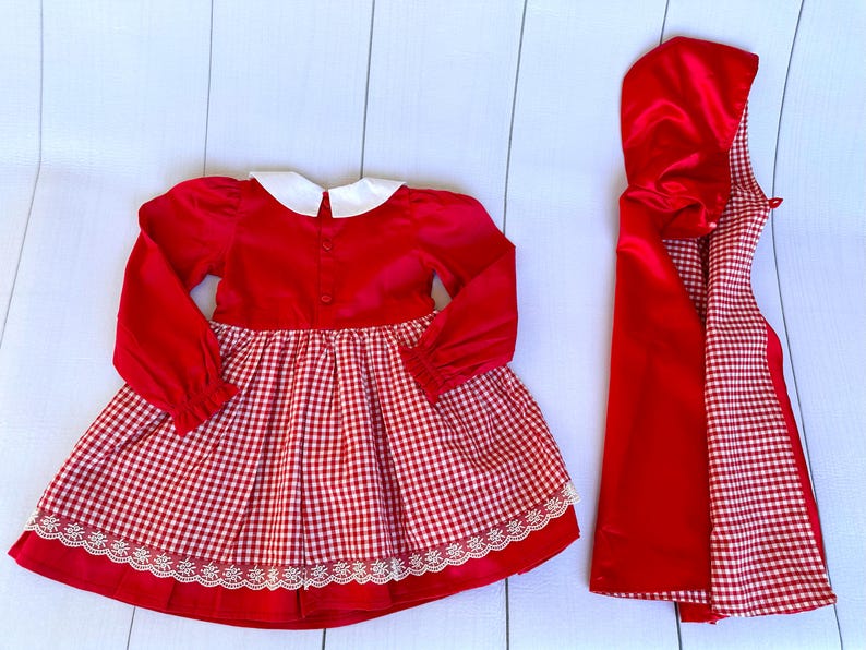 K&ouml;nnte beinhalten: Rotes und wei&szlig;es Gingham-Kleid mit wei&szlig;em Kragen und passender roter Kapuze. Das Kleid hat einen wei&szlig;en Spitzenbesatz.
