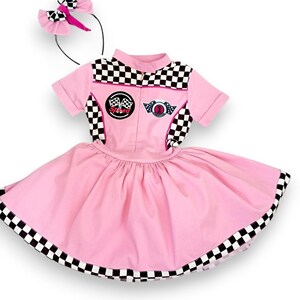 May include: Pink and black checkered racing-themed dress with a pink bow headband. The dress has a pink skirt with a black and white checkered trim and a black and white checkered top with a pink zipper and patches that say "Speed" and "Champion".