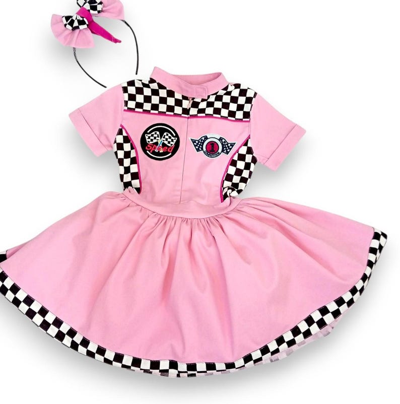 May include: Pink and black checkered racing-themed dress with a pink bow headband. The dress has a pink skirt with a black and white checkered trim and a black and white checkered top with a pink zipper and patches that say "Speed" and "Champion".