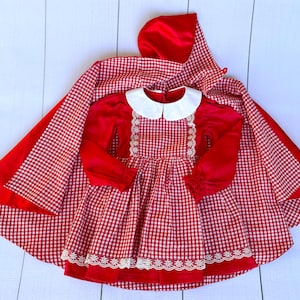 K&ouml;nnte beinhalten: Rotes und wei&szlig;es Gingham-Kleid mit wei&szlig;em Kragen und Spitzenbesatz. Das Kleid wird mit einem roten Satin-Umhang kombiniert.