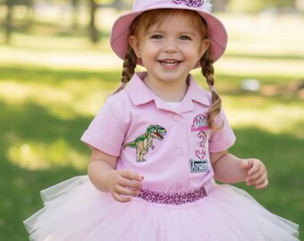 May include: A young child wearing a pink outfit with a dinosaur theme. The outfit includes a pink collared shirt with a dinosaur patch, a tutu skirt, and a matching hat with a bow. The shirt has the number 3 and the name "Annabelle" on it.