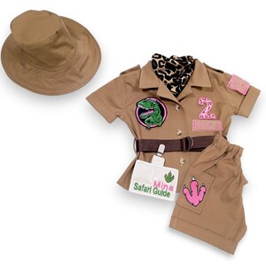 Puede incluir: Un conjunto de disfraz de safari color canela, que incluye una camisa de manga corta, pantalones y un sombrero de ala ancha. La camisa presenta un parche de dinosaurio, una etiqueta con el nombre "Mina Safari Guide" y un cintur&oacute;n. Los pantalones tienen un parche rosa con una huella de dinosaurio.