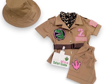 Puede incluir: Un conjunto de disfraz de safari color canela, que incluye una camisa de manga corta, pantalones y un sombrero de ala ancha. La camisa presenta un parche de dinosaurio, una etiqueta con el nombre "Mina Safari Guide" y un cinturón. Los pantalones tienen un parche rosa con una huella de dinosaurio.
