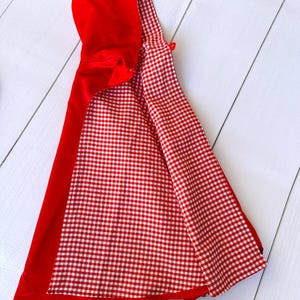 K&ouml;nnte beinhalten: Rote Kapuze mit wei&szlig;-rotem Karomuster als Futter. Die Kapuze ist gefaltet und liegt flach auf einer wei&szlig;en Holzoberfl&auml;che.
