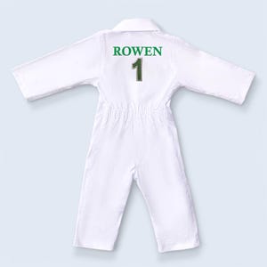 Macro view of the green thread embroidery on the white fabric, displaying the name "ROWEN 1". The image highlights the density of the stitches and the vibrant green color that stands out against the white cotton. It serves as a testament to the craftsmanship and durability of the personalized elements of the outfit.