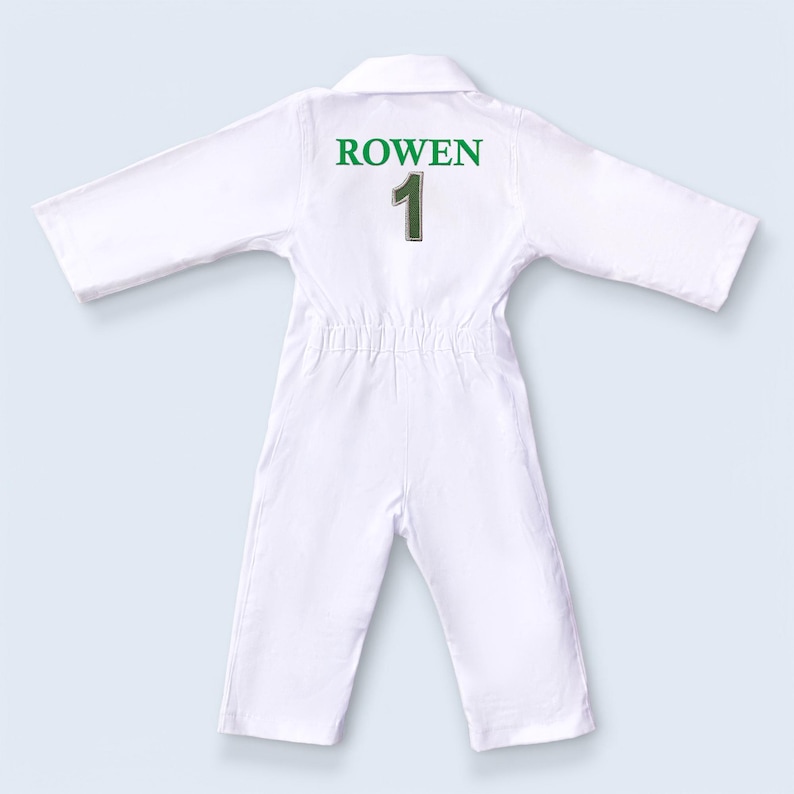 Macro view of the green thread embroidery on the white fabric, displaying the name "ROWEN 1". The image highlights the density of the stitches and the vibrant green color that stands out against the white cotton. It serves as a testament to the craftsmanship and durability of the personalized elements of the outfit.