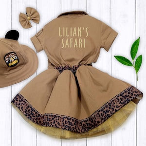 May include: A brown safari-themed dress with a leopard print trim and a matching hat with black ears. The dress has the text "Lillian's Safari" on the back.