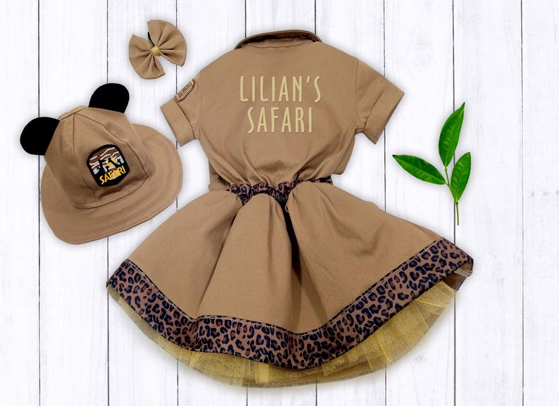 May include: A brown safari-themed dress with a leopard print trim and a matching hat with black ears. The dress has the text "Lillian's Safari" on the back.