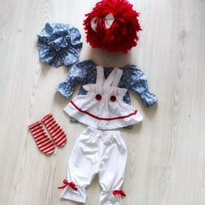 May include: A doll outfit set featuring a blue floral print bonnet, a red yarn wig with white bows, a blue floral print top with white suspenders, a white skirt with red trim, white bloomers with red bows, and red and white striped socks.