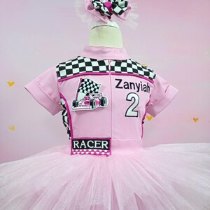 May include: Pink tutu dress with a black and white checkered racing top. The top has a pink racing car graphic, the text "Zanyiah 2", and the word "RACER".