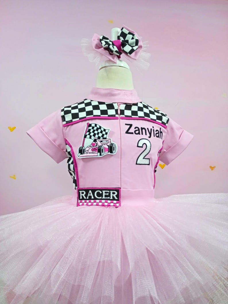 May include: Pink tutu dress with a black and white checkered racing top. The top has a pink racing car graphic, the text "Zanyiah 2", and the word "RACER".
