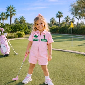May include: A young person in a pink golf-themed jumpsuit with the name "Chloe" on the front, holding a pink golf club. The outfit has a "Green One" patch. A pink golf bag and clubs are in the background on a green golf course.