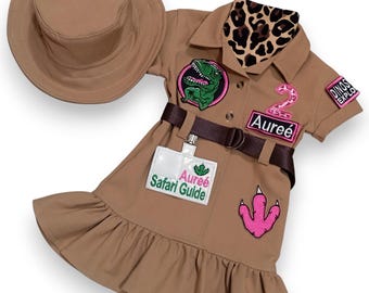 May include: A tan safari-style outfit for a child, including a dress with a belt, patches, and a matching hat. The dress features a dinosaur patch, a pink number 2, and the text "Safari Guide". A leopard print scarf is attached.