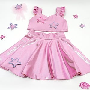 May include: A pink two-piece outfit for a girl, featuring a crop top with ruffle sleeves and a skirt. The top has a sweetheart neckline and is decorated with purple and pink starfish appliques. The skirt is a full circle skirt with a shiny, iridescent finish.