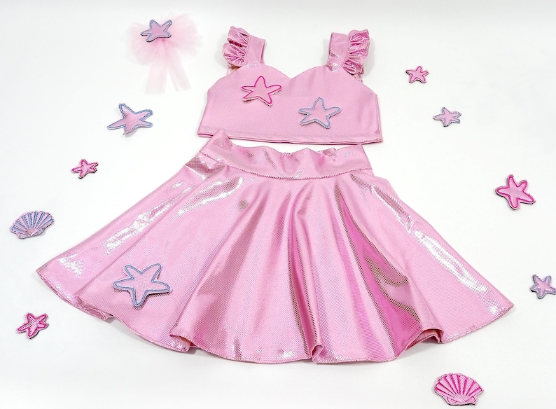 May include: A pink two-piece outfit for a girl, featuring a crop top with ruffle sleeves and a skirt. The top has a sweetheart neckline and is decorated with purple and pink starfish appliques. The skirt is a full circle skirt with a shiny, iridescent finish.