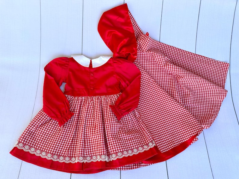 K&ouml;nnte beinhalten: Rot-wei&szlig;es Gingham-Kleid mit rotem Samt-Oberteil und wei&szlig;em Kragen. Das Kleid hat einen wei&szlig;en Spitzenbesatz und wird mit einer passenden roten Samt-Kapuzenjacke geliefert.