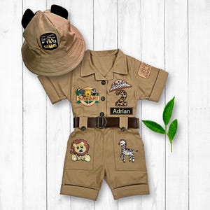May include: A tan safari-themed outfit for a toddler, featuring a safari hat with ears, a short-sleeved jumpsuit with safari patches, a belt, and a name tag that says "Adrian".