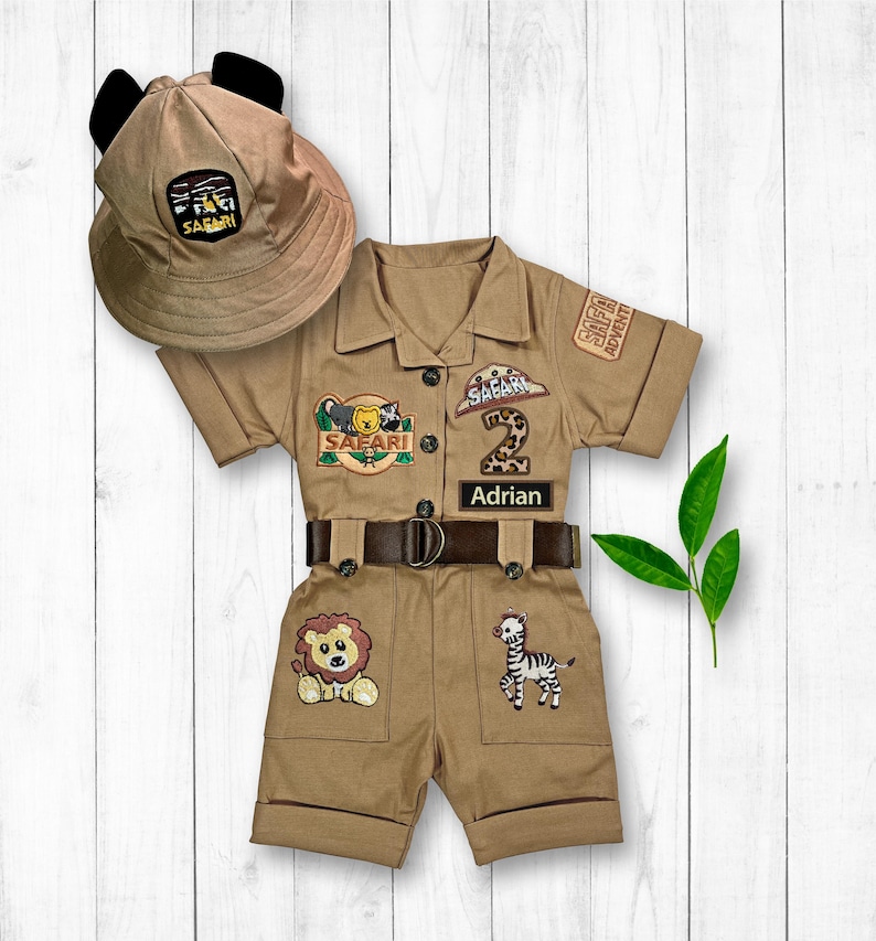 May include: A tan safari-themed outfit for a toddler, featuring a safari hat with ears, a short-sleeved jumpsuit with safari patches, a belt, and a name tag that says "Adrian".