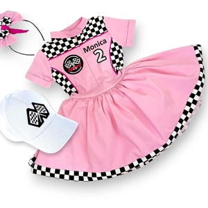 May include: Pink racing-themed dress with black and white checkered trim, a white cap with a checkered flag design, and a black headband with a pink bow. The dress has the name "Monica" and the number "2" on the front.
