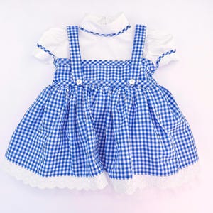 May include: A white short-sleeved dress featuring a blue and white gingham pinafore overlay with two shoulder straps, two white buttons at the waist, and a white scalloped lace trim along the bottom hem of the skirt and the collar neckline.