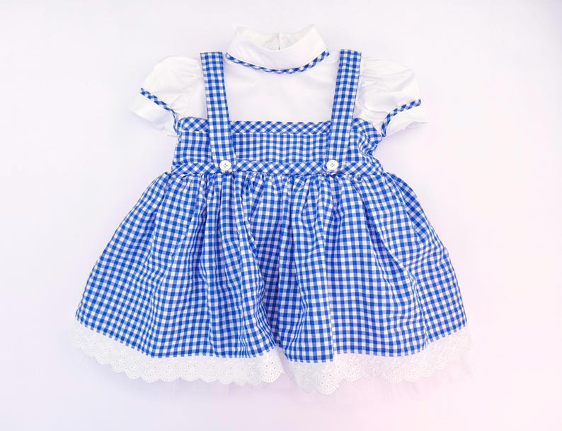 May include: A white short-sleeved dress featuring a blue and white gingham pinafore overlay with two shoulder straps, two white buttons at the waist, and a white scalloped lace trim along the bottom hem of the skirt and the collar neckline.