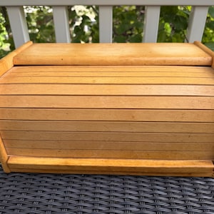 May include: A light brown wooden bread box featuring a curved roll-top lid with horizontal slats. The rectangular container has a smooth finish and a solid base, designed with a rounded frame on both sides to accommodate the sliding door mechanism.