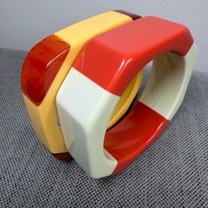 May include: Two vintage, chunky bangles. One is yellow with a dark red accent, the other is red and white. The bracelets are made of a glossy, hard material. The bangles are stacked together.