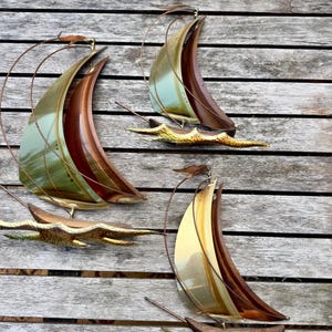 May include: Three wall-mounted metal sculptures of sailboats. Each piece features a polished brass sail, a dark brown curved mast, and a textured, gold-toned wave base. The metal sails have a smooth, reflective finish with subtle green and gold hues.