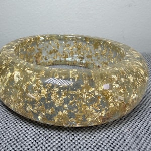 May include: A clear, round bracelet with gold leaf flakes embedded throughout. The bracelet has a thick, chunky design and a smooth, polished surface. The gold leaf adds a touch of luxury and sparkle to the accessory.