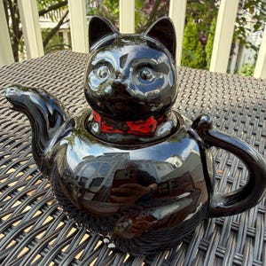 Op de afbeelding: Een glanzend zwarte keramische theepot in de vorm van een kattenkop, compleet met oren en een rode vlinderdas. De theepot heeft een gebogen tuit en handvat en staat op een geweven oppervlak.