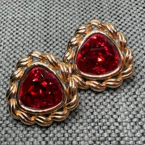 May include: A pair of gold-toned earrings featuring a triangular, faceted red stone set within a twisted rope-style metal border. The design showcases a polished metallic finish and a vibrant, deep red center that reflects light across its surface.