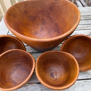 May include: A collection of wooden bowls in varying sizes. The largest bowl is in the background, with four smaller bowls in front. The bowls are a warm brown color, with visible wood grain patterns. The bowls are likely for serving food.