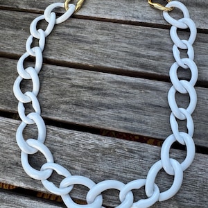 May include: A white oval link necklace with a gold-tone chain and clasp. The necklace is made of large, glossy white links. The gold chain is connected to the white links on each side. The necklace is displayed on a wooden surface.