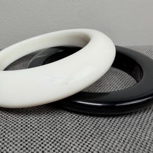 May include: Two oval bangles, one white and one black, rest on a textured gray surface. The white bangle is positioned on top of the black one, showcasing their contrasting colors and smooth, rounded shapes. Jewelry.
