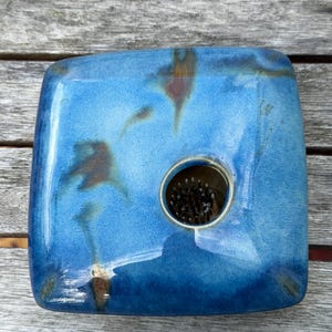 May include: A square ceramic flower frog with a glossy blue glaze featuring dark brown mottled accents. The center contains a circular opening with a metal pin holder, known as a kenzan, featuring numerous sharp, upright metal needles for arranging flowers.