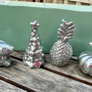 May include: A set of four polished silver-toned metal figurines including a rabbit, a Christmas tree with a star topper, a pineapple, and a pumpkin. The items are arranged in a row in front of a light green rectangular box.