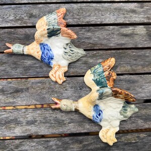 May include: Two ceramic wall-mounted mallard duck figures in flight. Each duck features a painted finish with a green head, a white neck ring, a blue speculum patch on the wings, and brown and tan plumage. The ducks are depicted with open beaks.