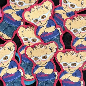 以下が含まれることがあります： サングラスをかけたクマの漫画のステッカーのコレクション。青いシャツには「OCCASIONALLY」の文字、ジーンズを着用。ステッカーは赤い縁取りがあり、黒い表面に配置されています。