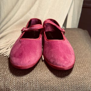 Pode incluir: Um par de sapatos baixos de veludo rosa com uma tira e um fecho de fivela.