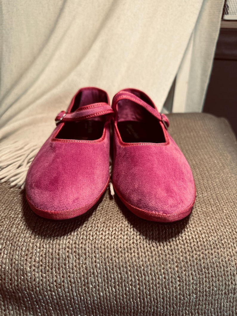 Pode incluir: Um par de sapatos baixos de veludo rosa com uma tira e um fecho de fivela.