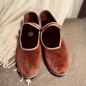 Puede incluir: Un par de bailarinas de terciopelo color &oacute;xido con ribete blanco. Los zapatos est&aacute;n sobre una superficie marr&oacute;n texturizada.