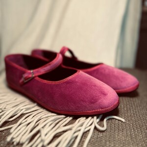 Pode incluir: Um par de sapatilhas de ballet de veludo rosa com uma tira e uma fivela. As sapatilhas est&atilde;o sobre uma superf&iacute;cie branca e fofa.