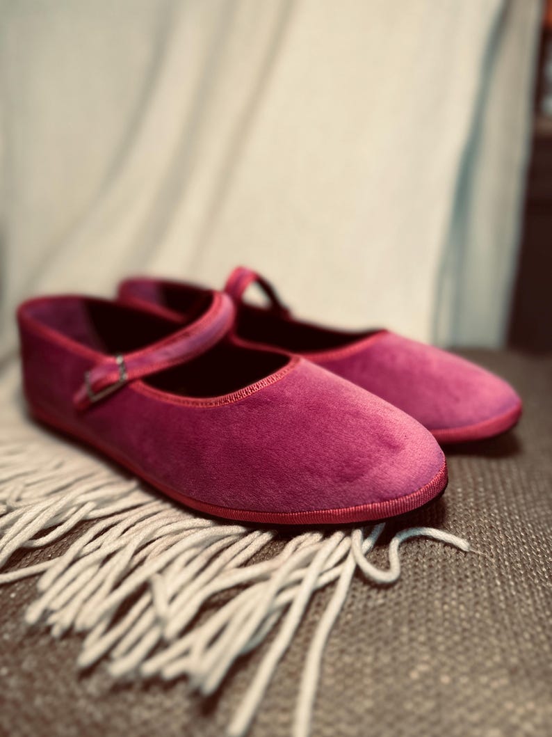 Pode incluir: Um par de sapatilhas de ballet de veludo rosa com uma tira e uma fivela. As sapatilhas est&atilde;o sobre uma superf&iacute;cie branca e fofa.