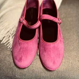 Pode incluir: Um par de sapatos de veludo rosa com uma tira e uma fivela. Os sapatos est&atilde;o sobre uma superf&iacute;cie texturizada.