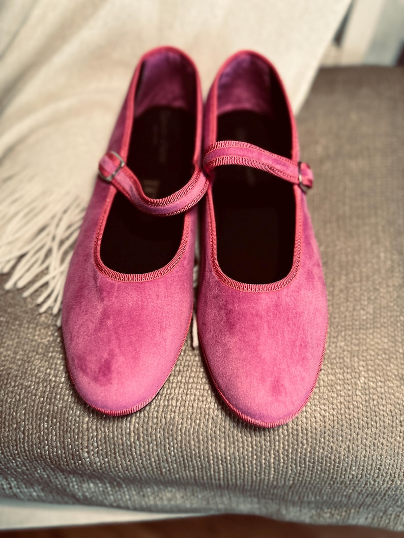 Pode incluir: Um par de sapatos de veludo rosa com uma tira e uma fivela. Os sapatos est&atilde;o sobre uma superf&iacute;cie texturizada.