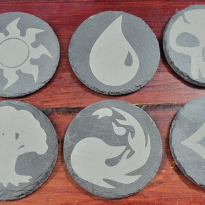 May include: Six round slate coasters with etched designs. Each coaster features a different symbol, including a sun, water droplet, skull, tree, flame, and diamond. The coasters are gray and sit on a wooden surface.