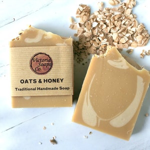 Puede incluir: Dos barras de jabón artesanal con el texto "OATS & HONEY" y "Traditional Handmade Soap". El jabón es de color marrón claro con un diseño en espiral y está envuelto en una etiqueta de papel marrón. Avena dispersa junto al jabón.