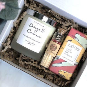 May include: A gift box filled with brown shredded paper contains a white candle with a silver lid, a small glass jar of matches, and a chocolate bar wrapped in colourful paper. The candle label reads "Orange & Cinnamon" and the chocolate bar label reads "COCO Salted Caramel Milk Chocolate".