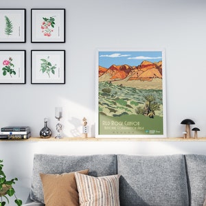 K&ouml;nnte beinhalten: Ein gerahmtes Poster mit einer farbenfrohen Illustration des Red Rock Canyon National Conservation Area in Nevada. Das Poster h&auml;ngt an einer wei&szlig;en Wand &uuml;ber einem Holzregal mit B&uuml;chern, Dekoartikeln und einer Topfpflanze.
