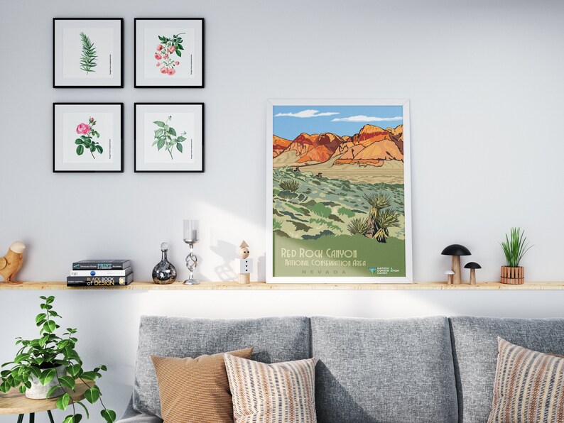K&ouml;nnte beinhalten: Ein gerahmtes Poster mit einer farbenfrohen Illustration des Red Rock Canyon National Conservation Area in Nevada. Das Poster h&auml;ngt an einer wei&szlig;en Wand &uuml;ber einem Holzregal mit B&uuml;chern, Dekoartikeln und einer Topfpflanze.